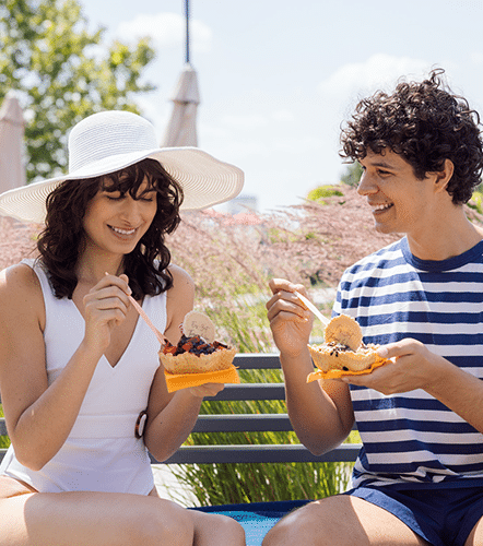 Junge Frau mit Sonnenhut und junger Mann essen entspannt in der Sonne ein Eis in einer Waffel und sitzen dabei auf einer Bank im Garten der Therme Wien.